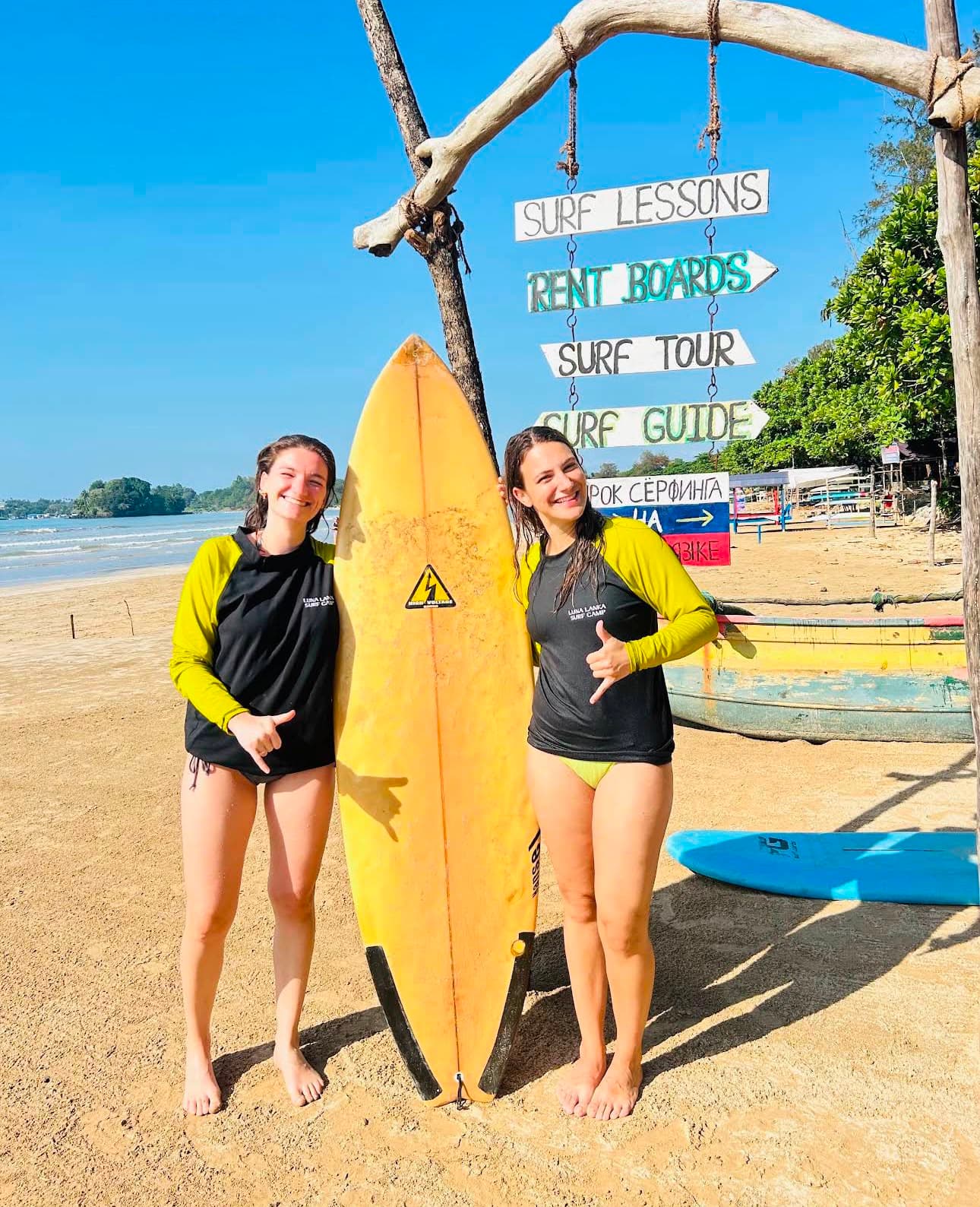 Two Luna Lanka surfers holding a surfboard beside the surf lessons sign